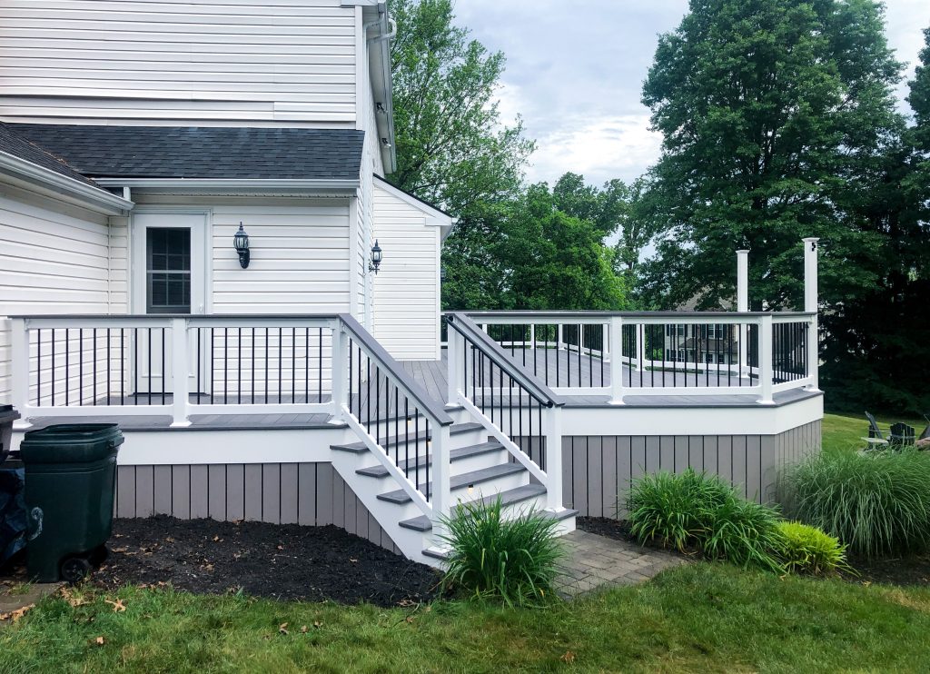 Left side view of a PVC deck with gray flooring, gray skirting, white railings, a white deck border, matching gray drink rails, a curved accent, and a matching staircase attached to a white house.