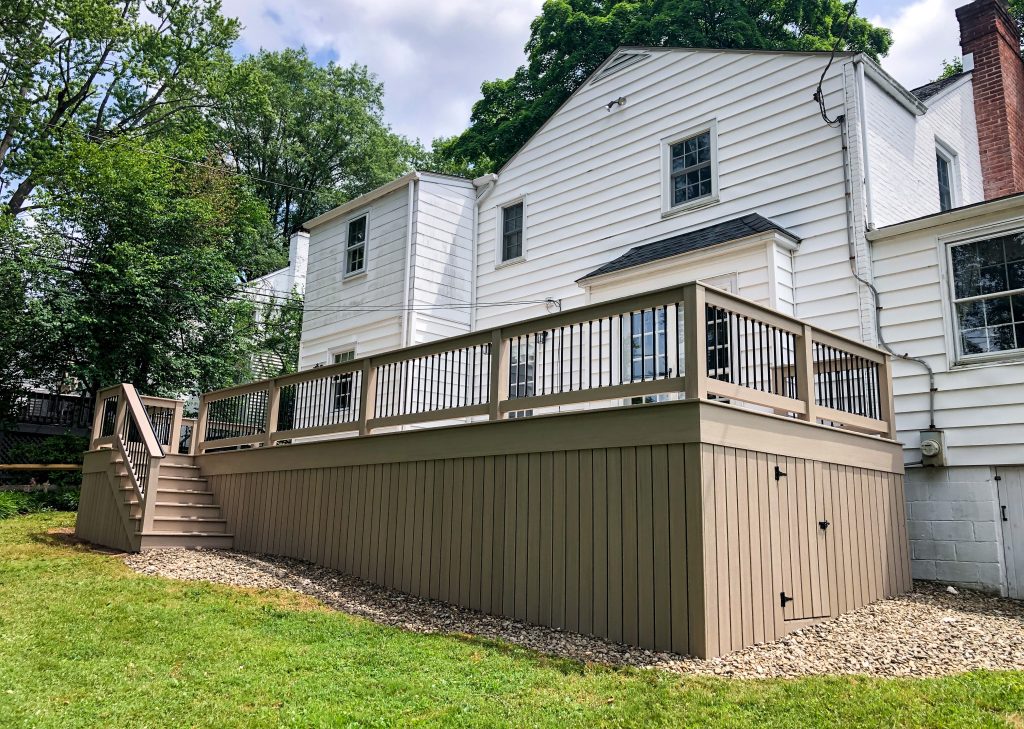 Front and right side view of a new PVC deck with light brown flooring, matching railings with black balusters, matching skirting with a door, and a staircase with black handrails attached to a white house.