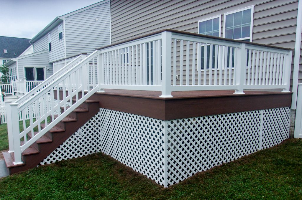 Front and right side view of a new dark brown PVC deck with vinyl lattice under deck, white vinyl railings, matching stairs, and a matching drink rail attached to a tan house.