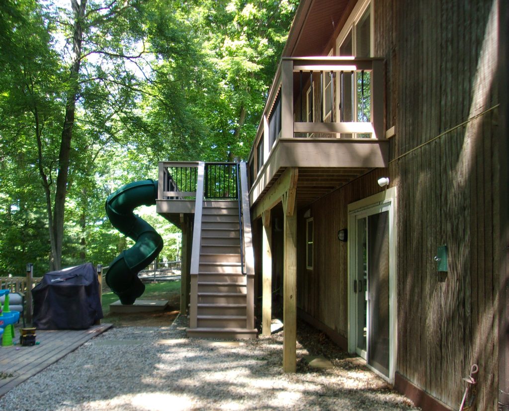 Ground side view of a second-story deck with light-colored PVC decking, matching railings, a matching staircase, and a green tunnel twist slide attached to the deck.