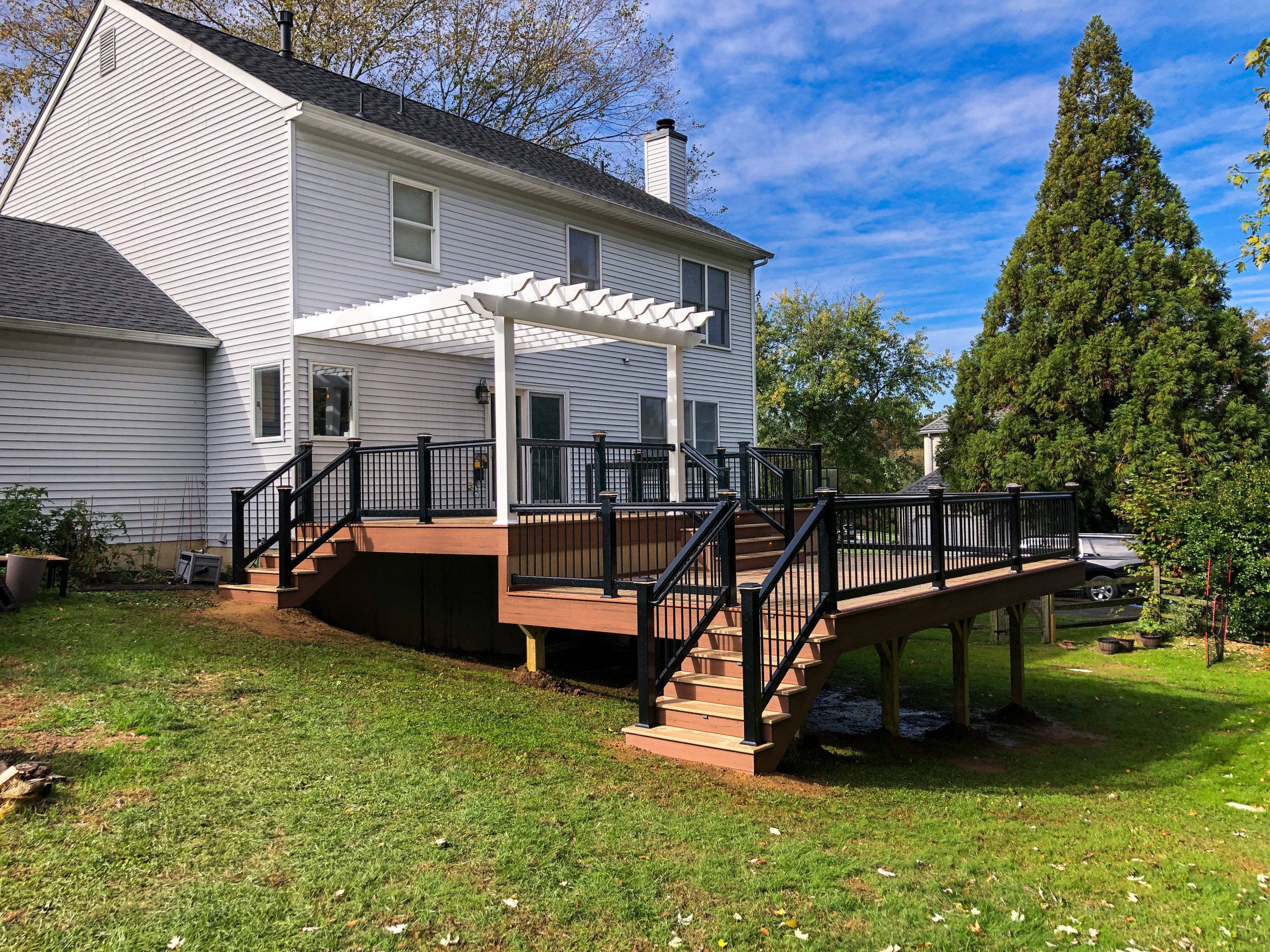 Front and left side view of a brown multi-level PVC deck with a dark brown deck border, 3 sets of matching stairs, a white pergola, and black railings with handrails.