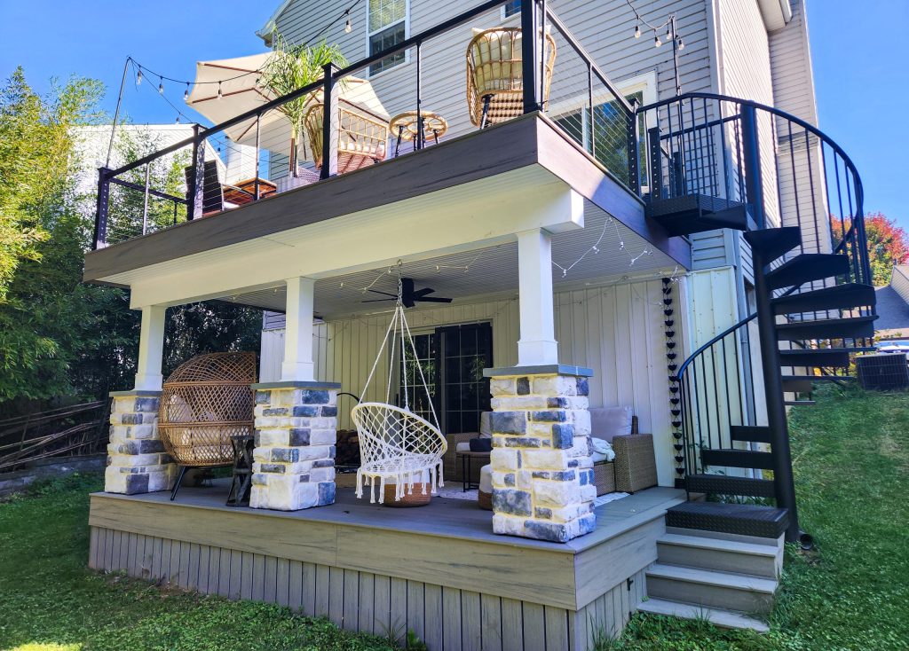 Front and right-side view of a 2 level deck with gray PVC decking, matching under-deck skirting, white trim, white posts, black railings with cable infill, a black spiral staircase, stone columns, and outdoor furniture.