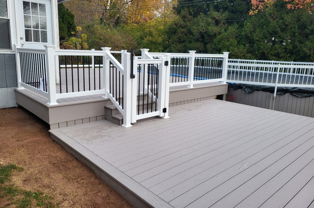 Gray PVC pool deck with 2 levels, white vinyl railings, black balusters, and a matching white gate with a code lock attached to a round above-ground pool.