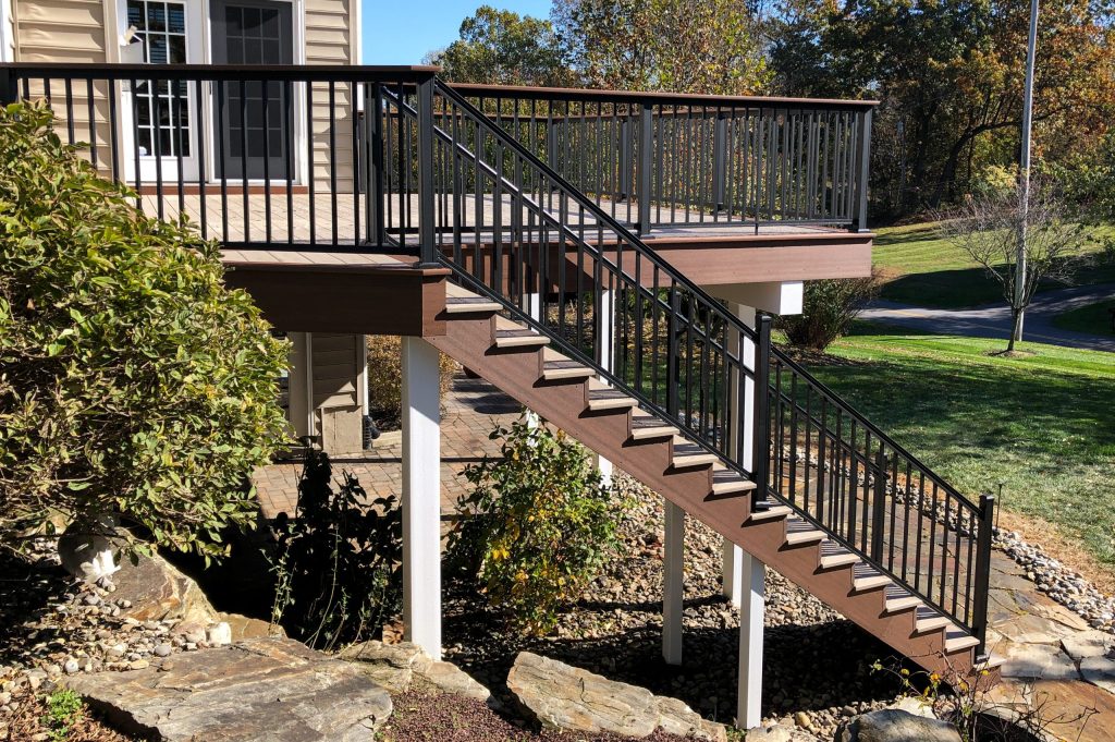 Side view of a tan second-story PVC deck with a brown border, matching drink rails, black aluminum railings, white posts, and a matching side staircase.